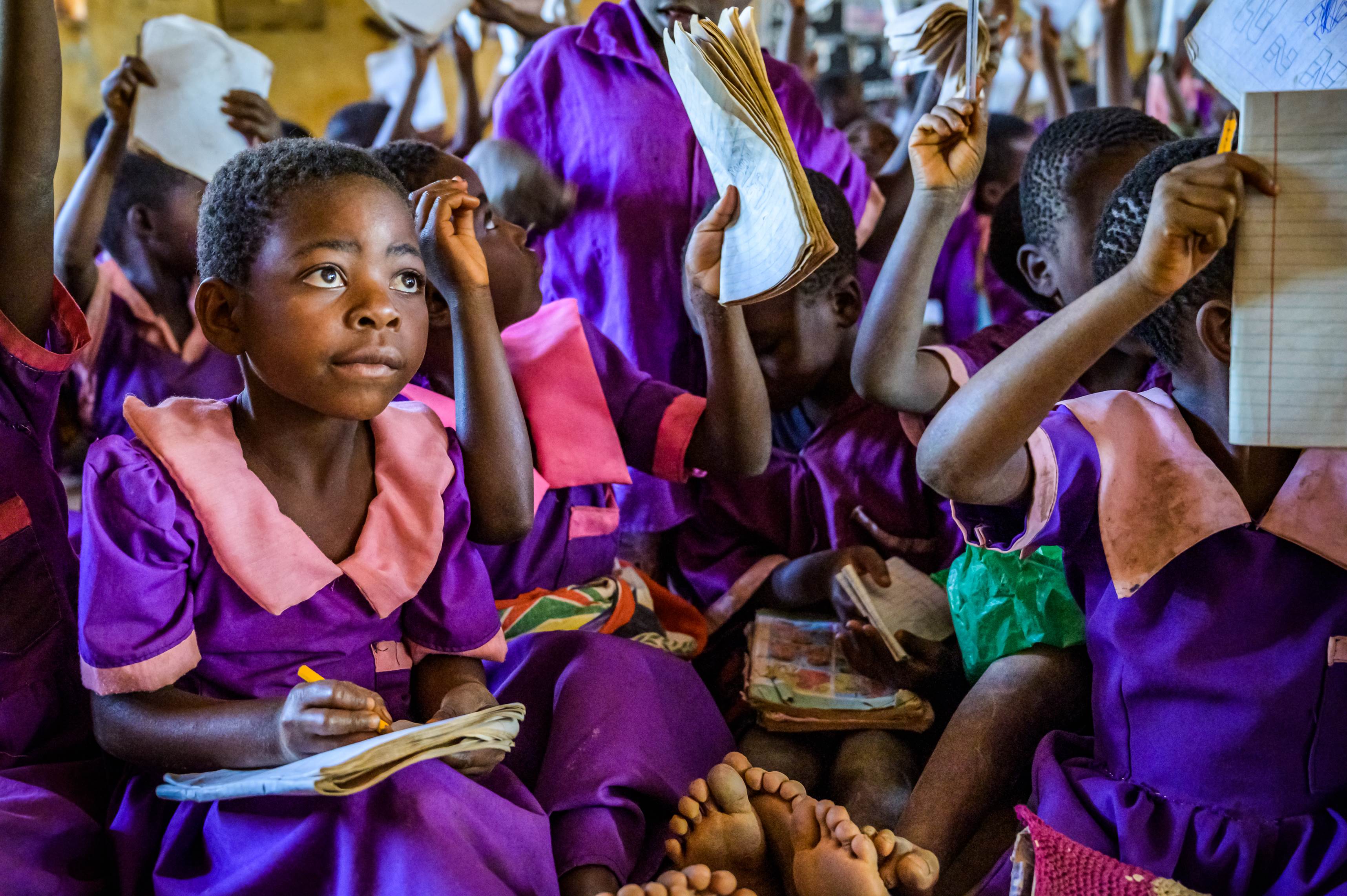 Una niña de Malawi en uniforme escolar con un cuaderno en su regazo escuchando la clase, con muchos compañeros detrás de ella