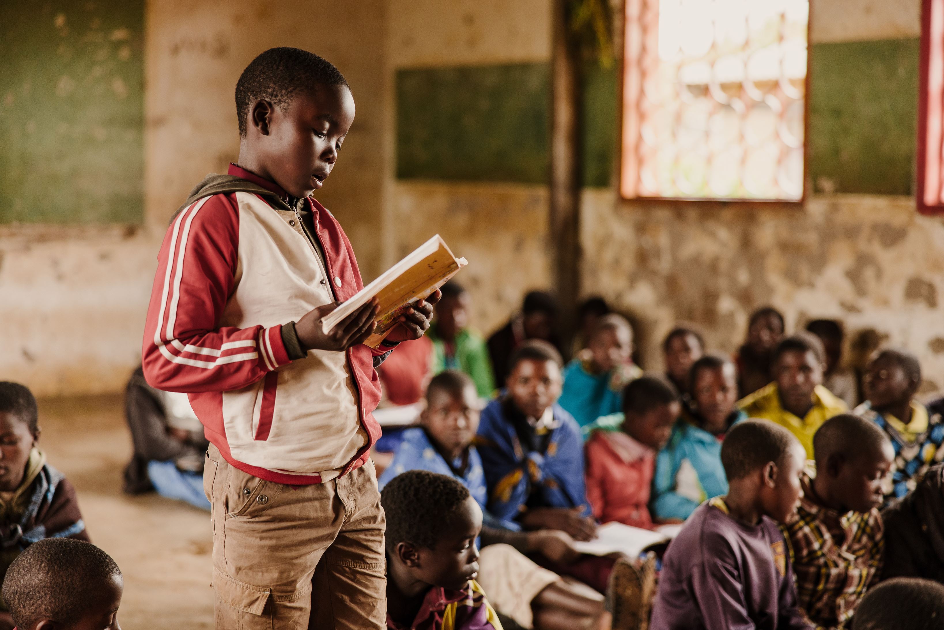 Niño de Malawi leyendo un libro de texto frente a un aula llena de alumnos sentados en el suelo.
