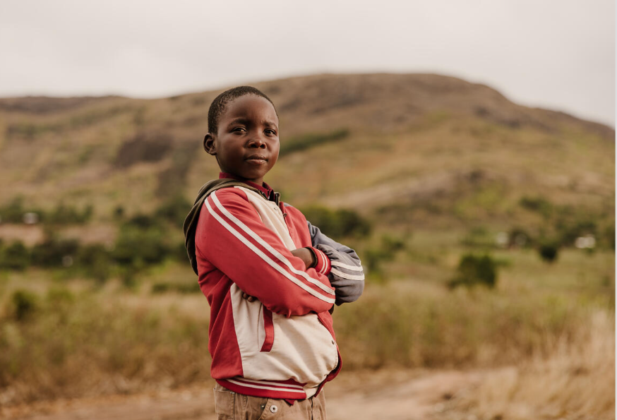 Niño de Malawi de pie con los brazos cruzados en un campo, mirando a la cámara