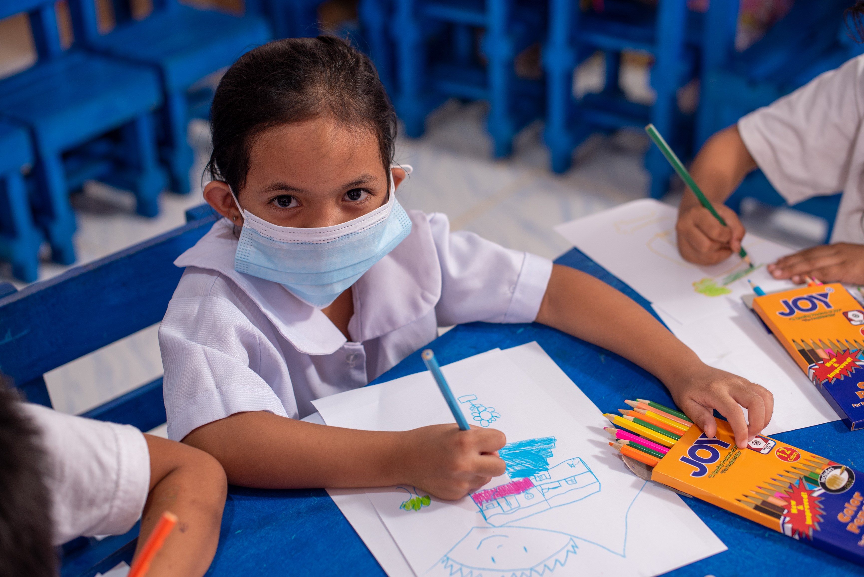 Niñas filipinas con mascarilla y uniforme escolar miran a la cámara mientras dibujan en la escuela