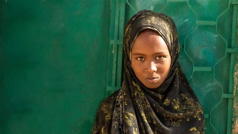 Chica de Bangladerh usando un hiyab, de pie frente a una puerta verde y mirando a la cámara