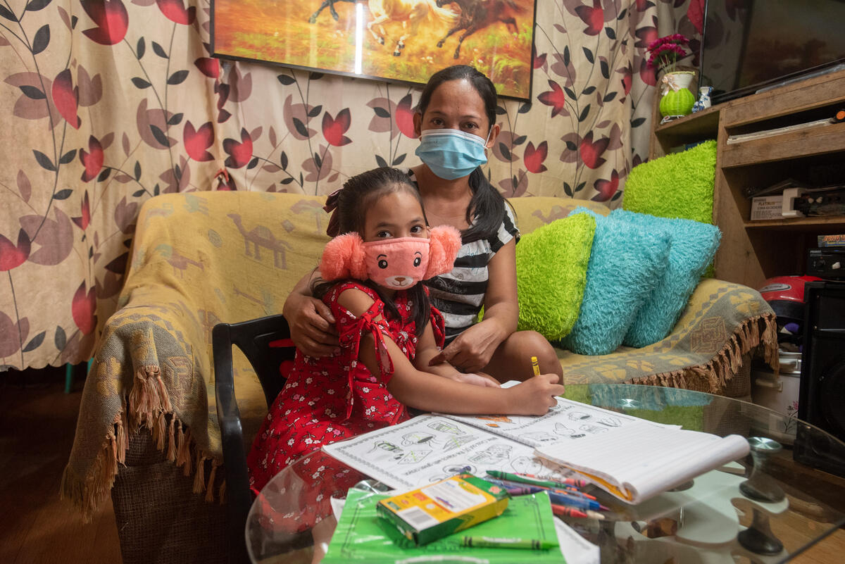 Niña filipina con una mascarilla rosa esponjosa sentada junto a su madre en una mesa, también con mascarilla.