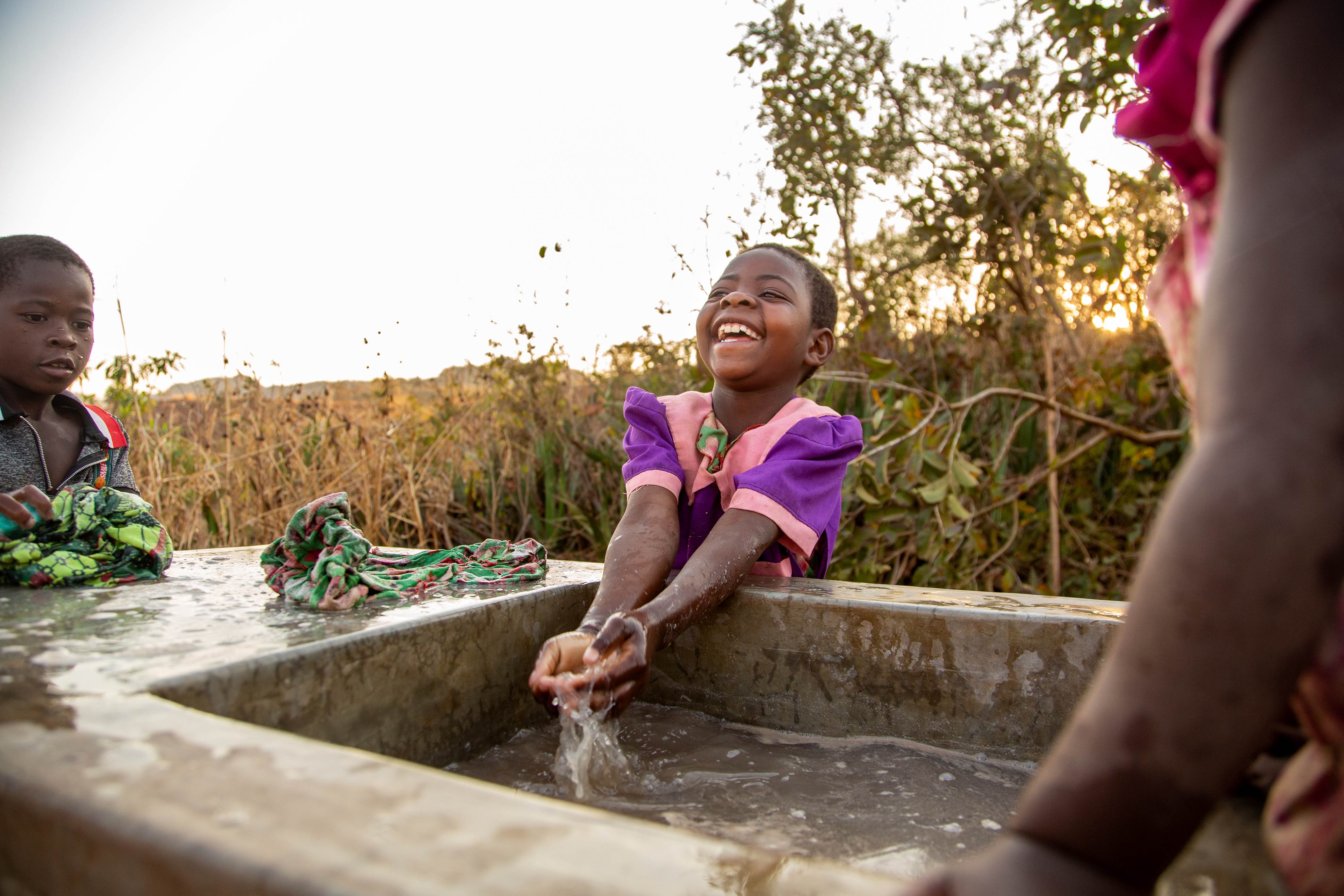 Niña de Malawi con un vestido morado brillante riendo mientras juega con el agua de un pozo local.