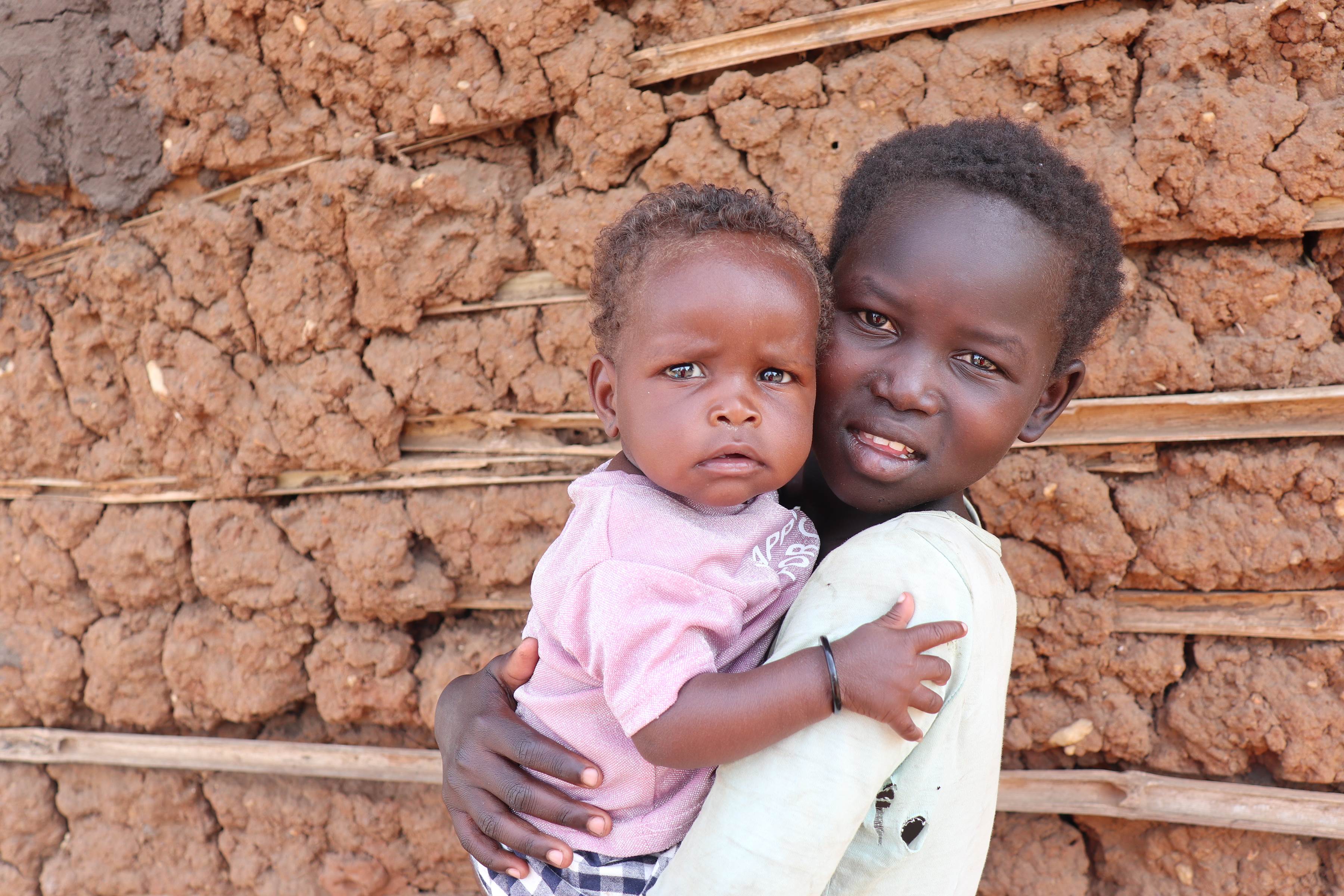 Un niño de ocho meses en Sudán del Sur abraza a un niño mayor sosteniéndolos, de pie frente a una pared