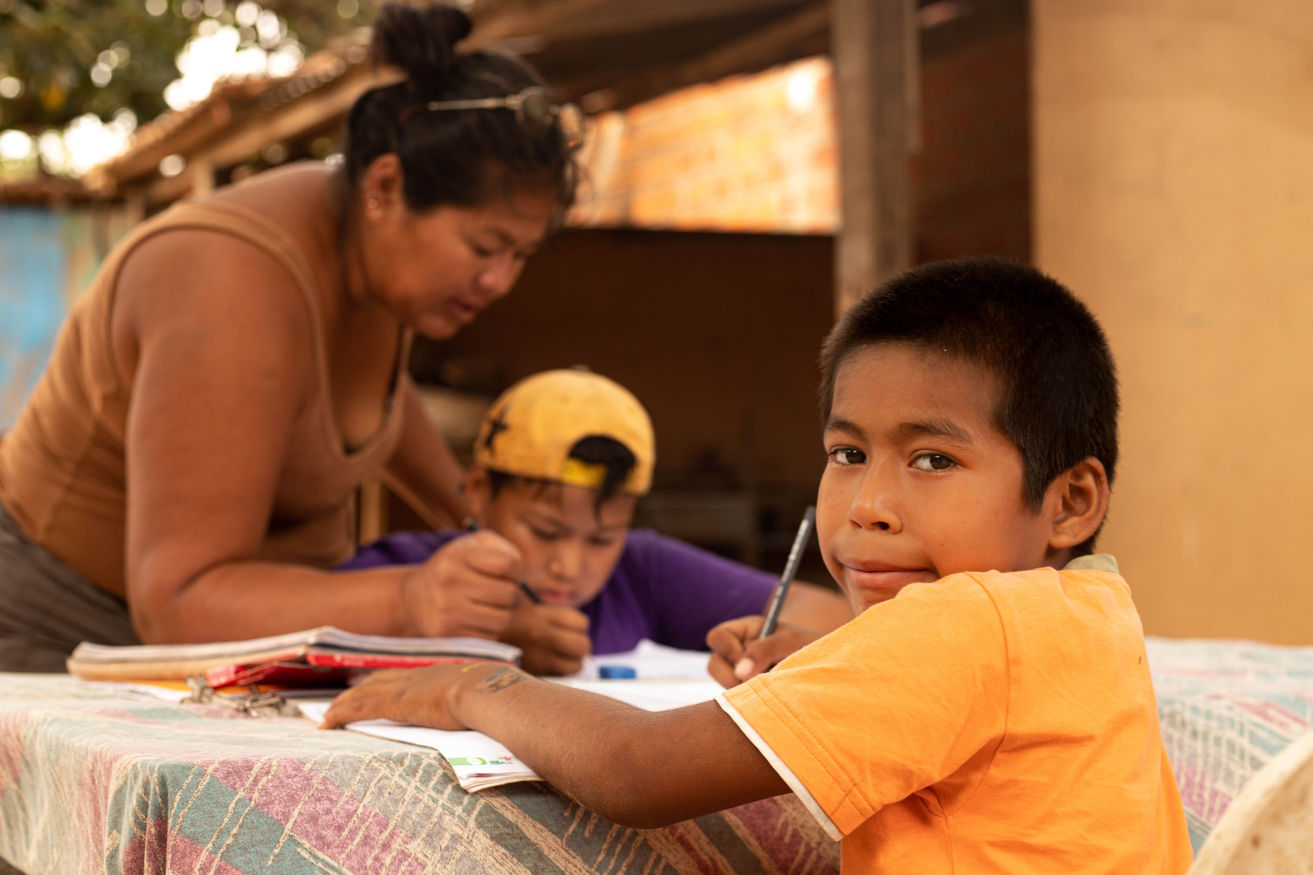 Niño de Bolivia que mira a la cámara mientras hace sus deberes. En el fondo se ve a una mujer y a un niño más joven escribiendo en un escritorio.