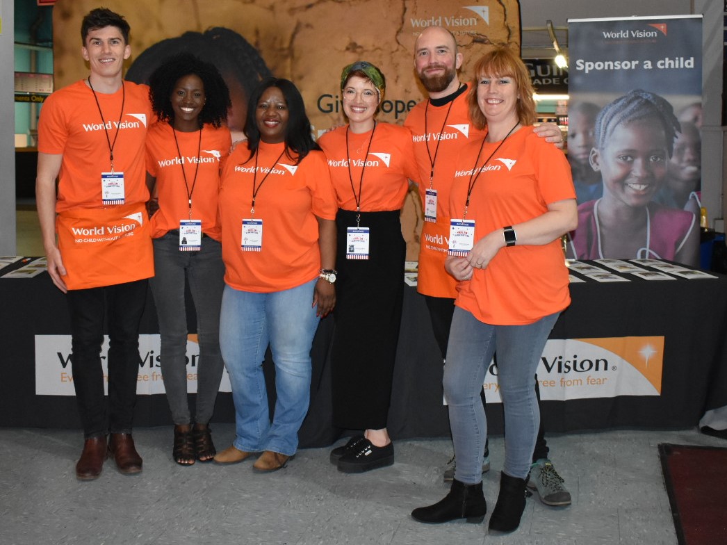 Seis miembros del personal de World Vision con camisetas naranjas frente a un puesto de voluntariado en un evento de World Vision