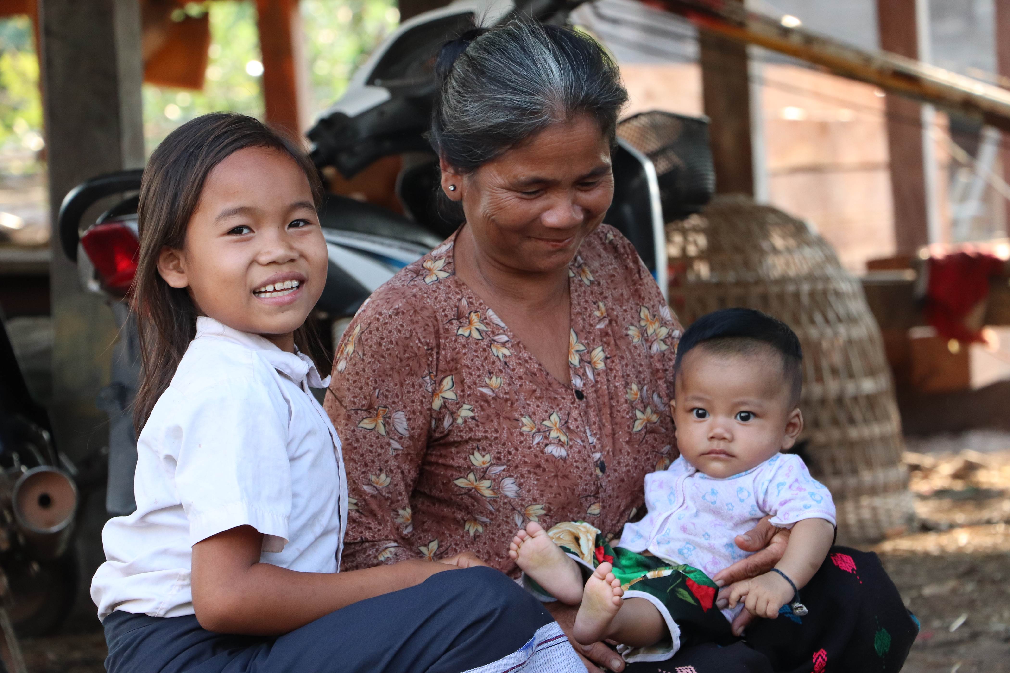 Niña de Laos sonriendo y mirando a la cámara junto a su abuela y su hermanita.