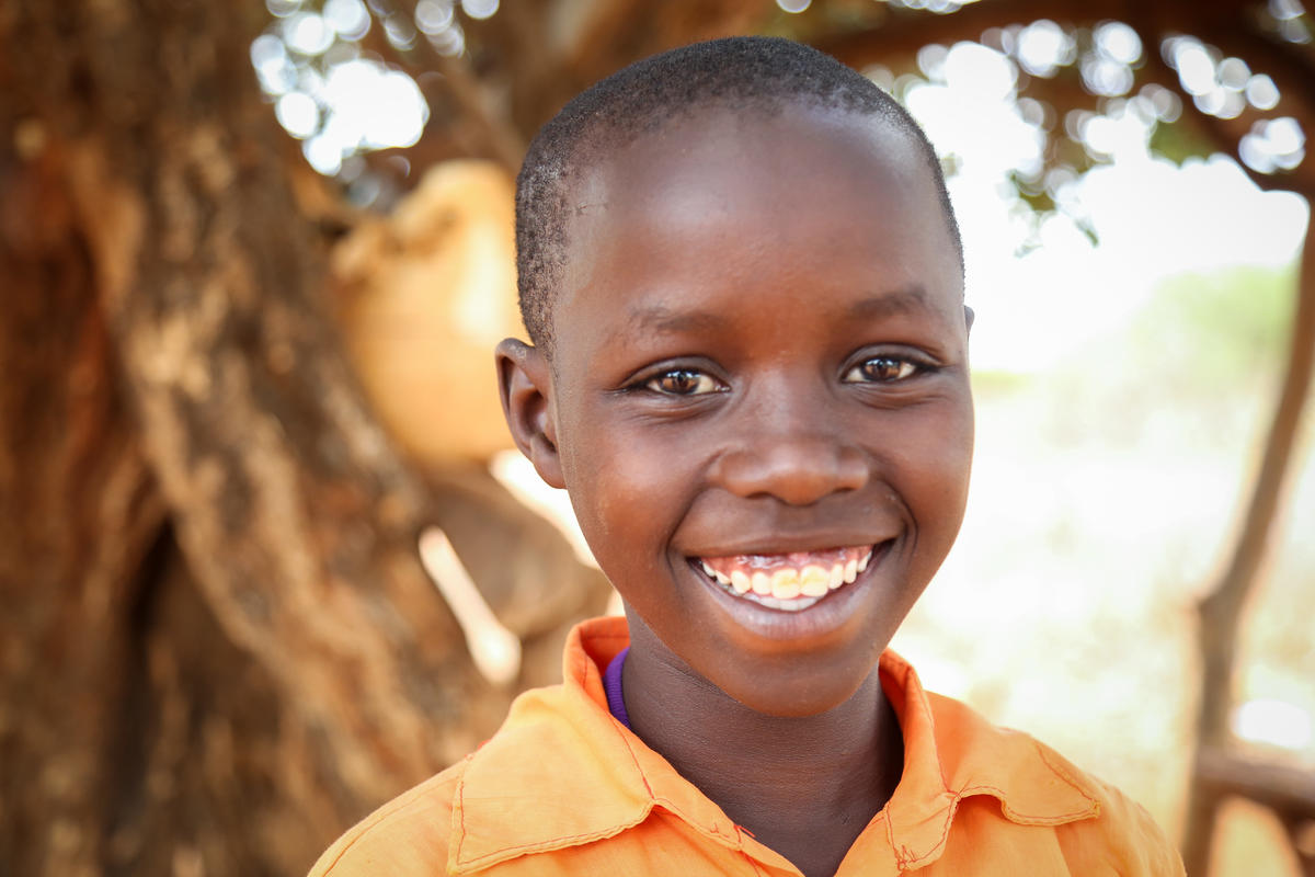 Niño keniano sonríe a la cámara, parado afuera frente a un árbol