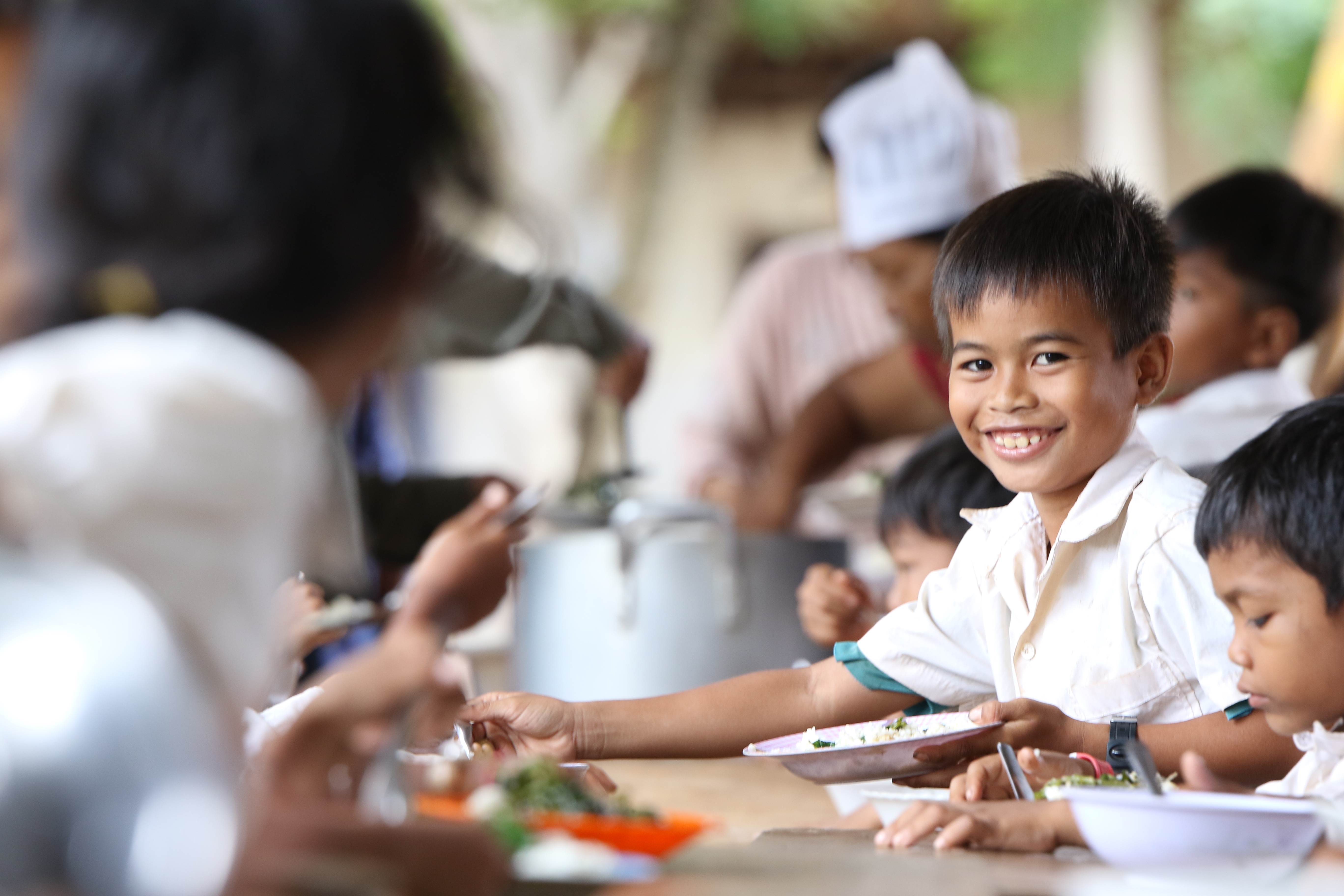Niño sonríe a la cámara, rodeado por sus compañeros de clase mientras comen su comida escolar gratuita en Camboya