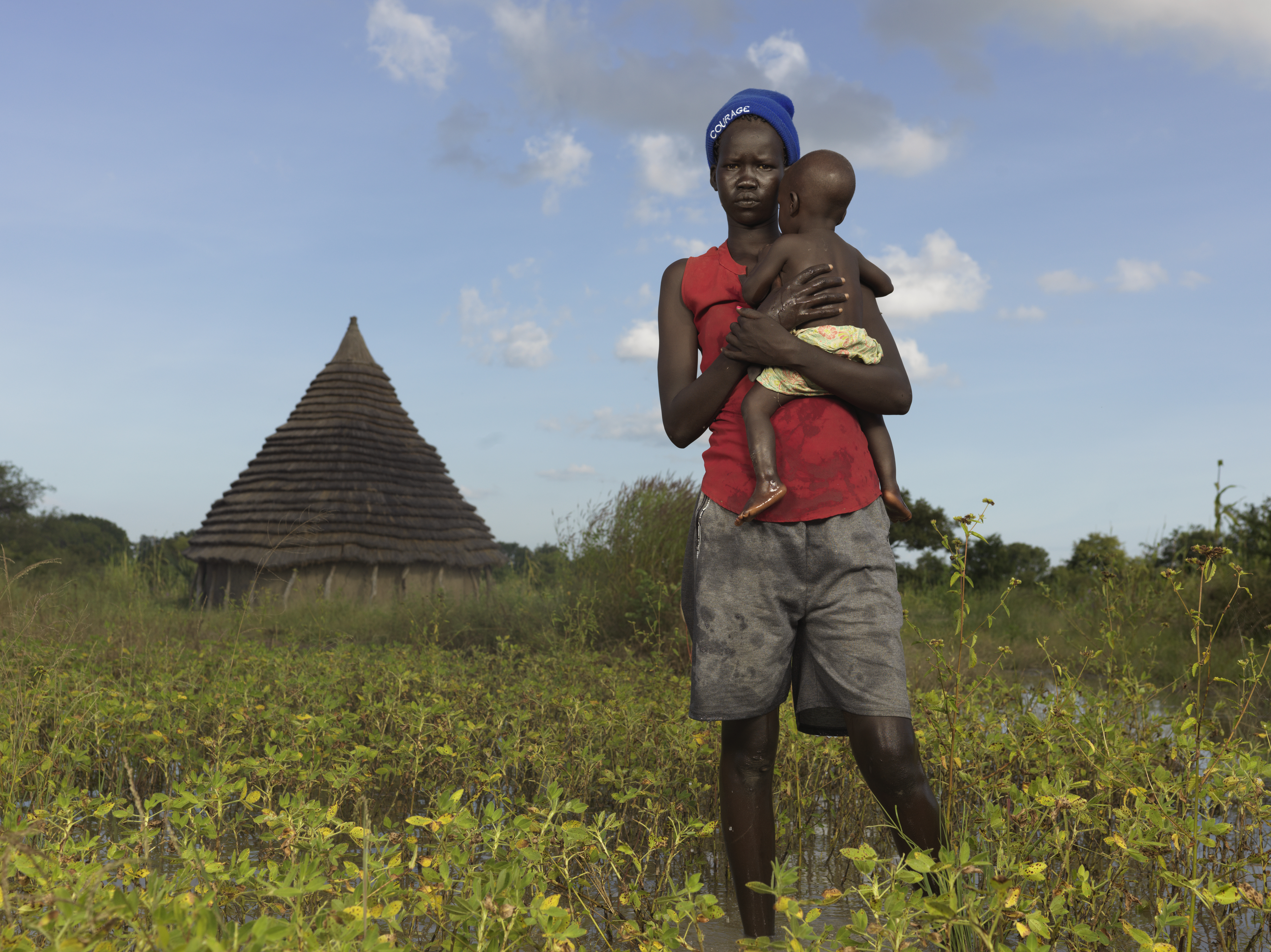 Madre adolescente de Sudán del Sur caminando por un campo mientras lleva a su hijo pequeño en brazos
