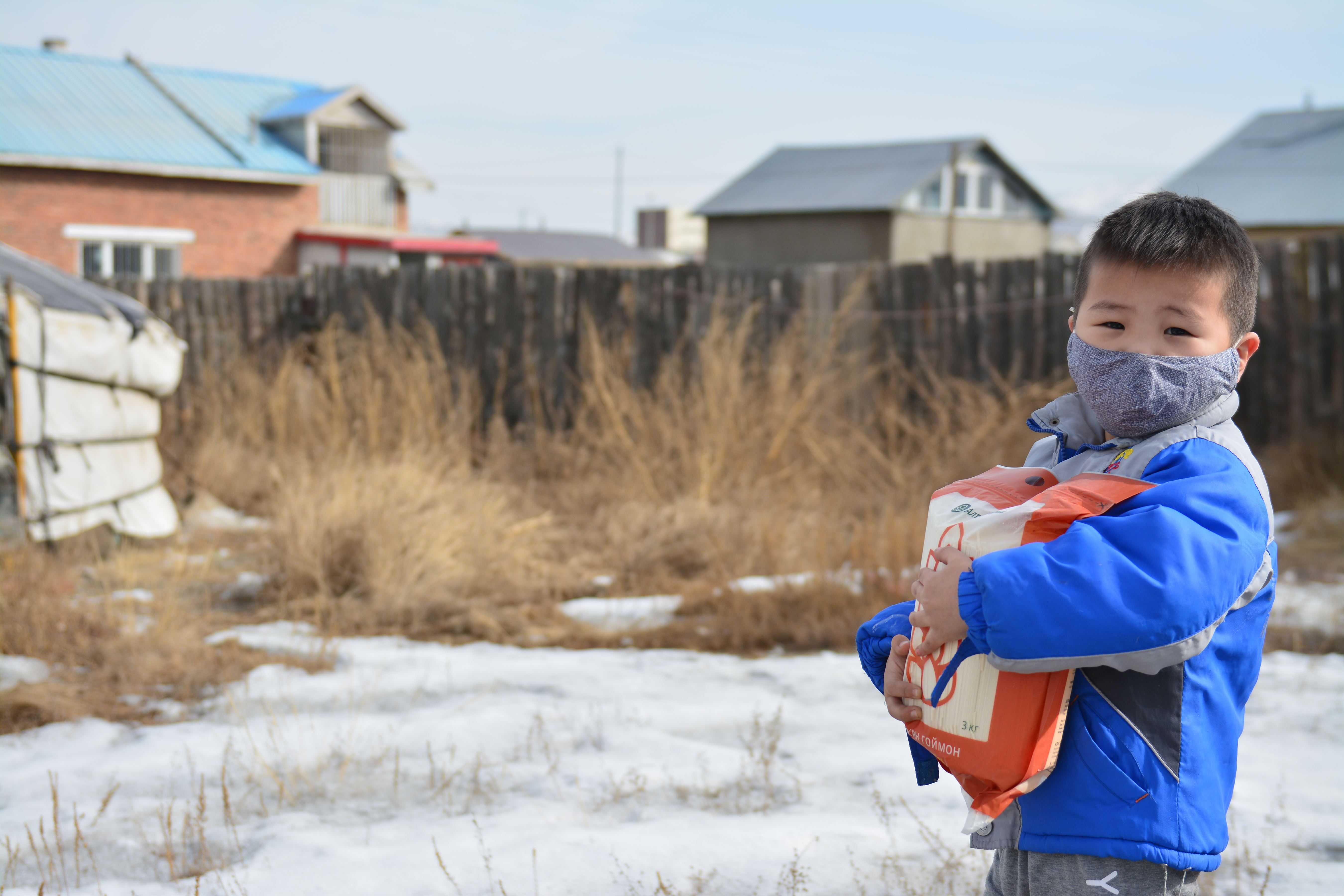 Un niño de Mongolia, con una mascarilla, lleva una bolsa de ayuda alimentaria a través de la nieve.