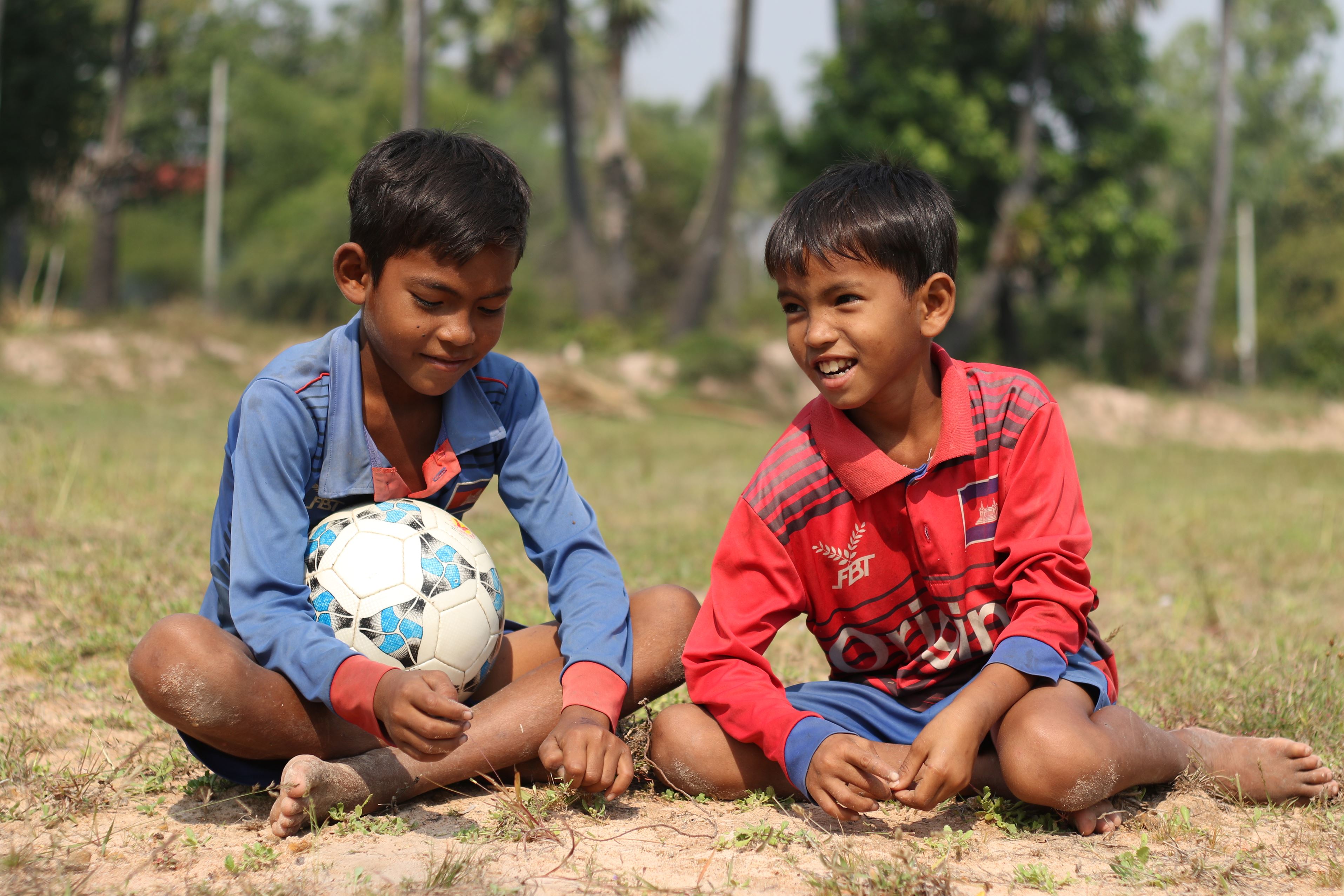 Dos niños sentados en un suelo de tierra, uno de ello tiene un balón de fútbol