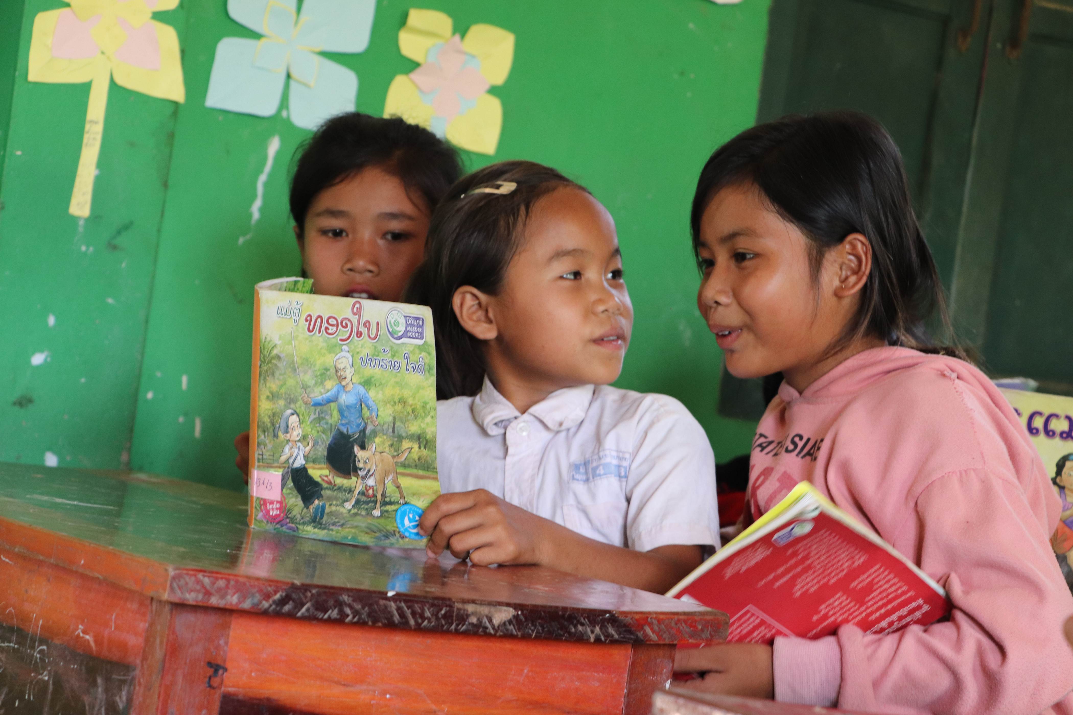 Tres escolares de Laos sentados en un pupitre y leyendo un libro infantil ilustrado.