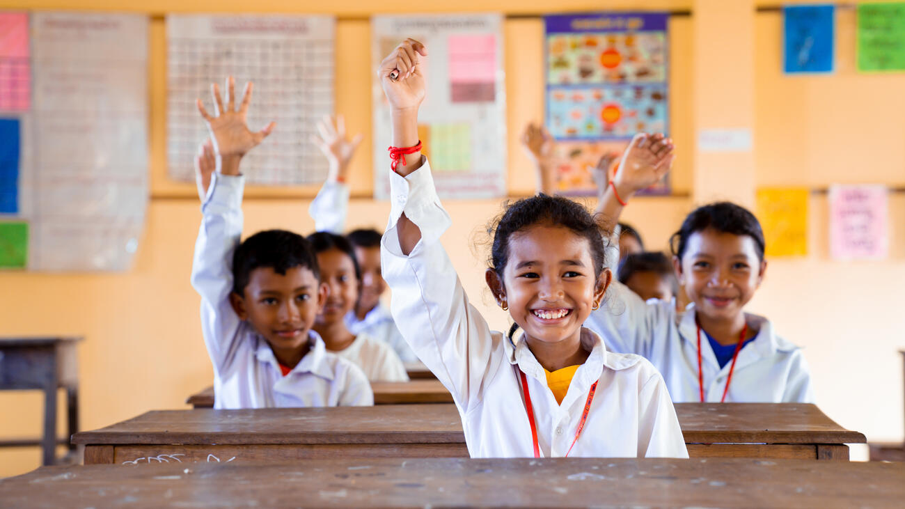 Niños camboyanos en una clase, todos con camisa blanca, levantando el brazo y sonriendo.