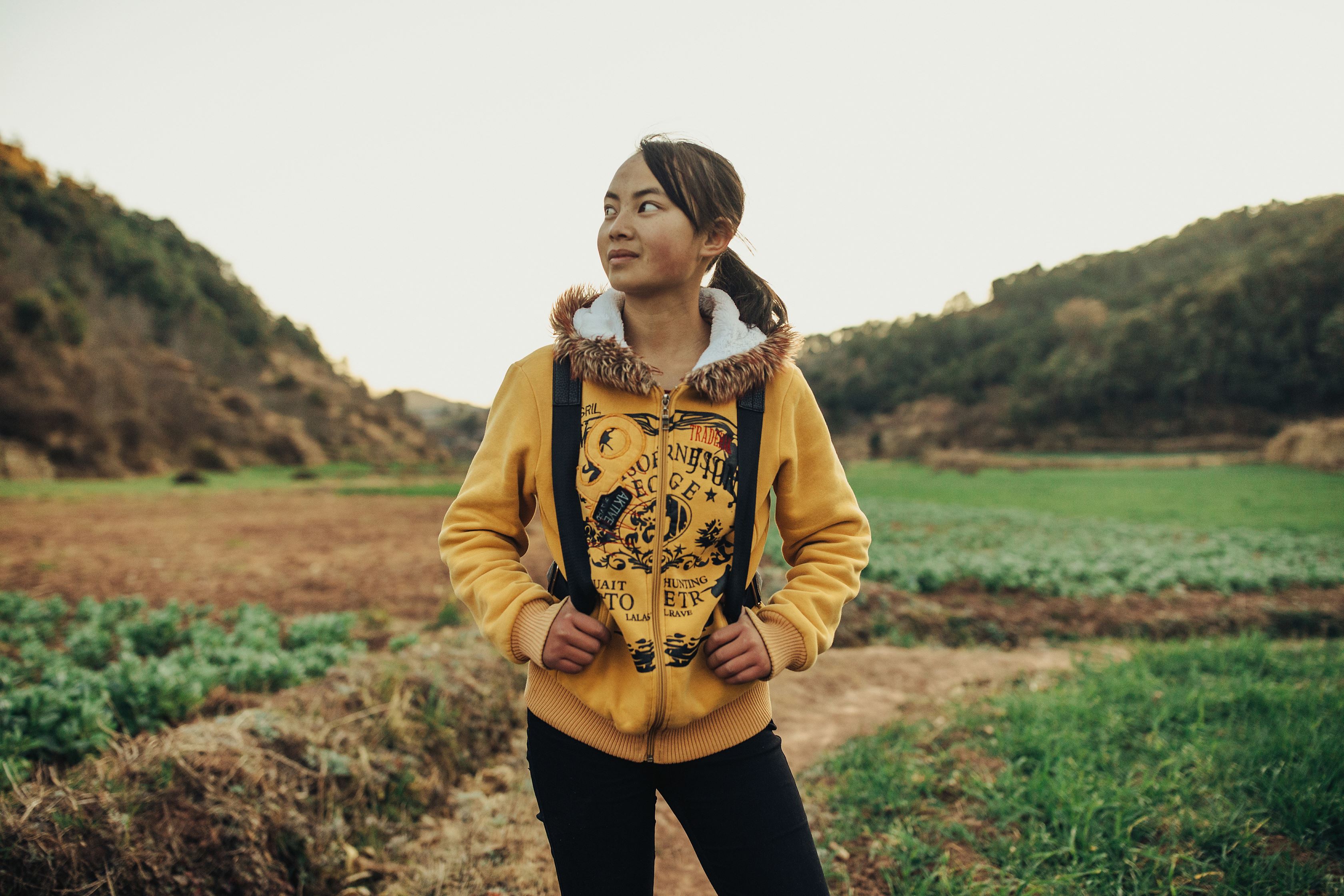 Niña china con capucha amarilla, llevando una mochila en un campo y mirando a lo lejos