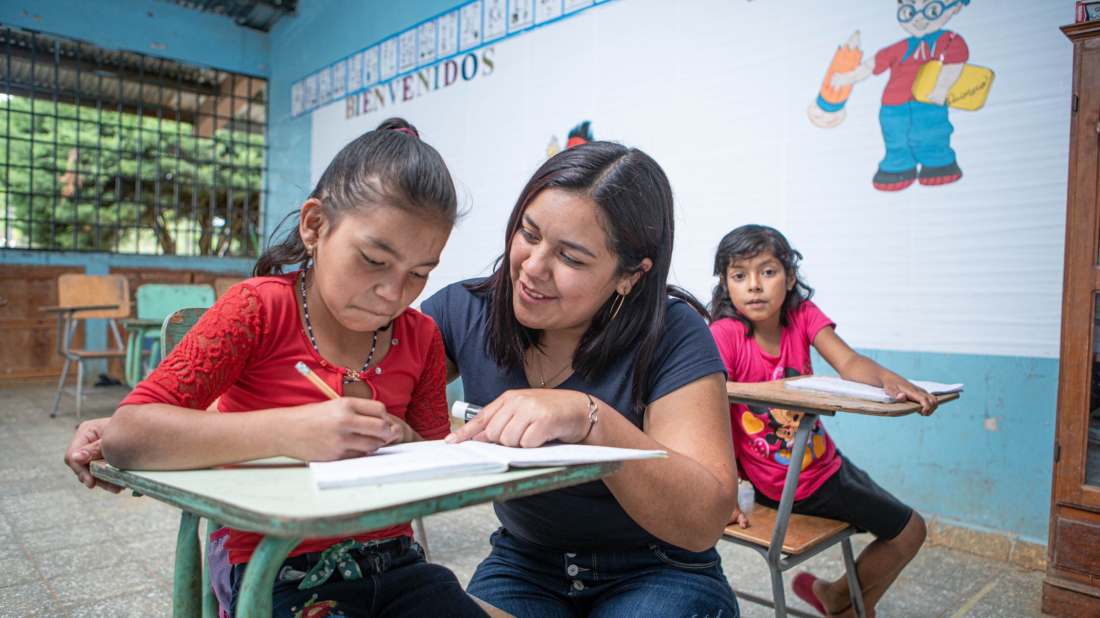 Profesora en Honduras ayudando a una alumna con su trabajo en su pupitre.