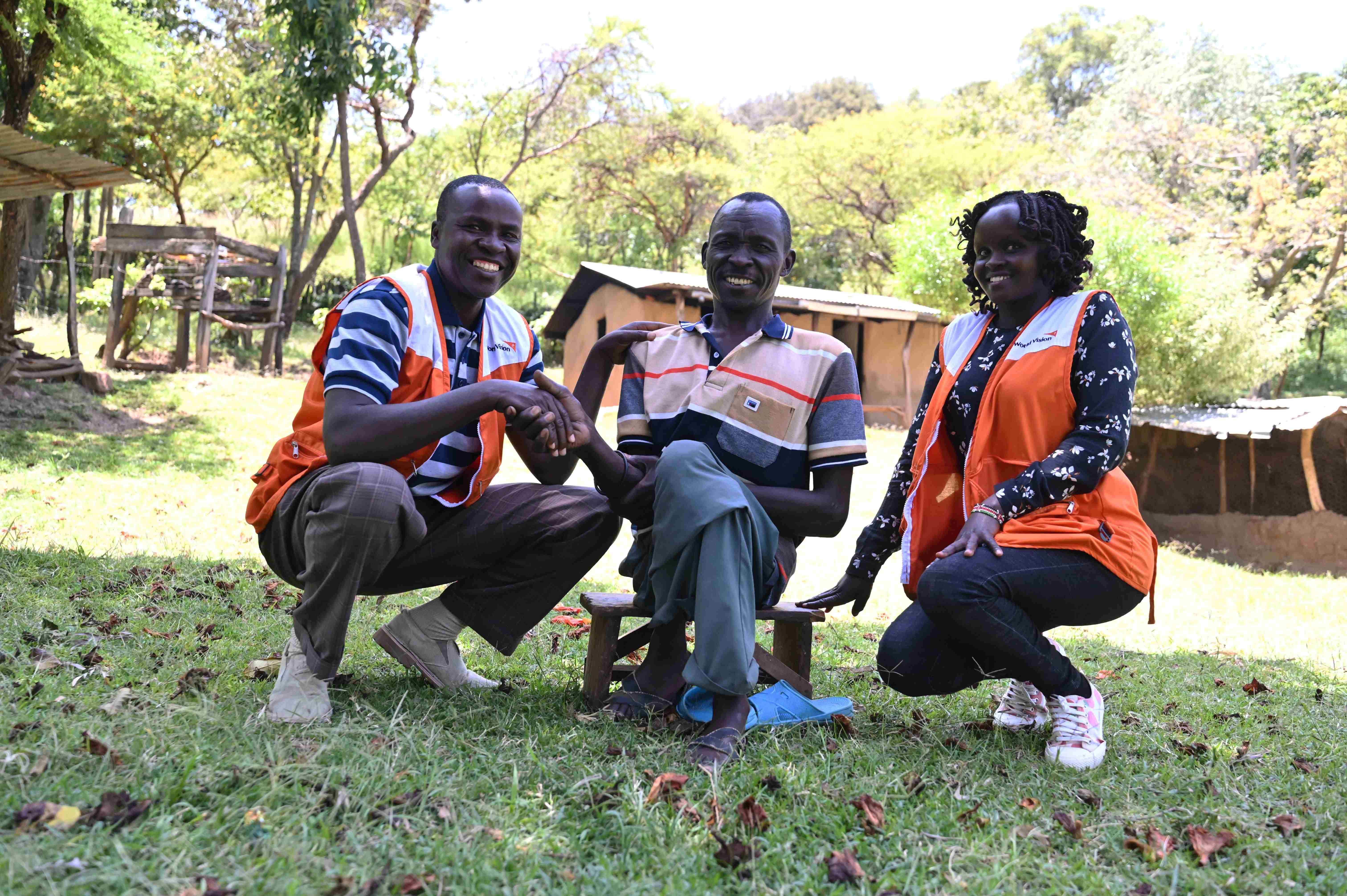 Farmer Samson sitting cross-legged in the middle of a field that he owns, smiling, with a World Vision representative on each side of him