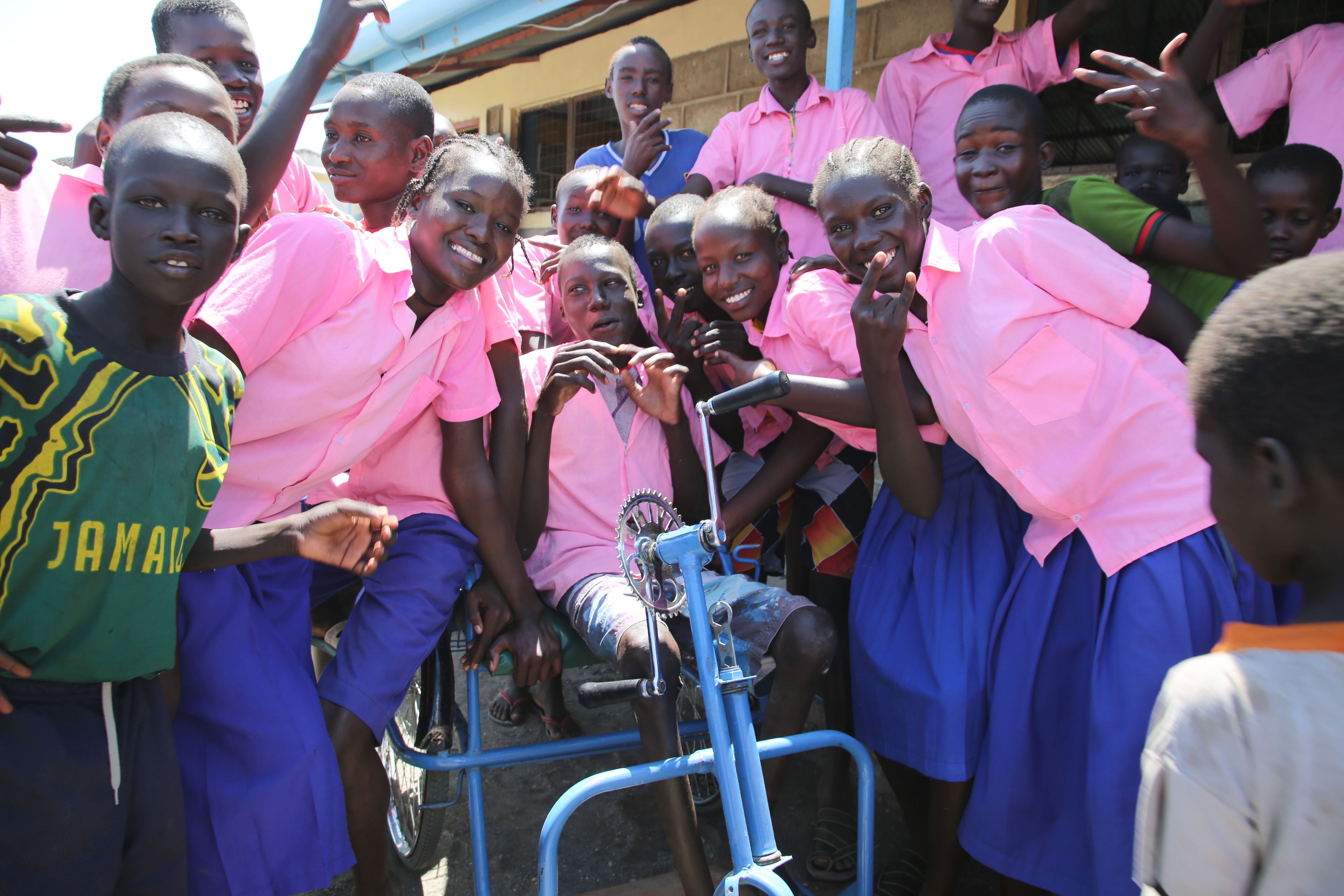 Un grupo de escolares de Uganda, vestidos con uniformes escolares rosas y azules, se reúnen en torno a su compañero de clase, sentado en una silla de ruedas en el centro del grupo.