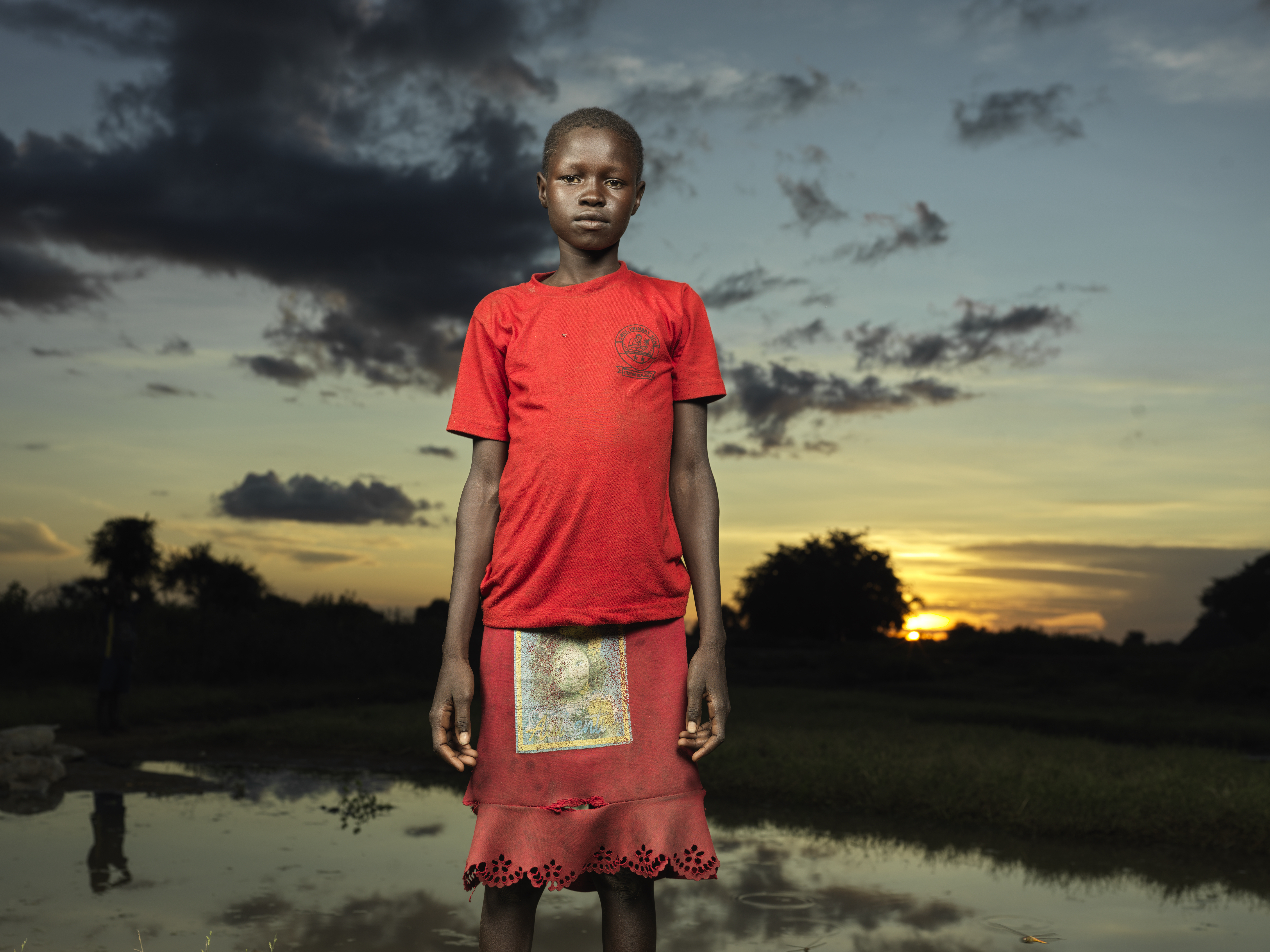 Niña de Sudán del Sur con un vestido rojo brillante fotografiada de pie y sola al atardecer en un campo de desplazados donde vive.