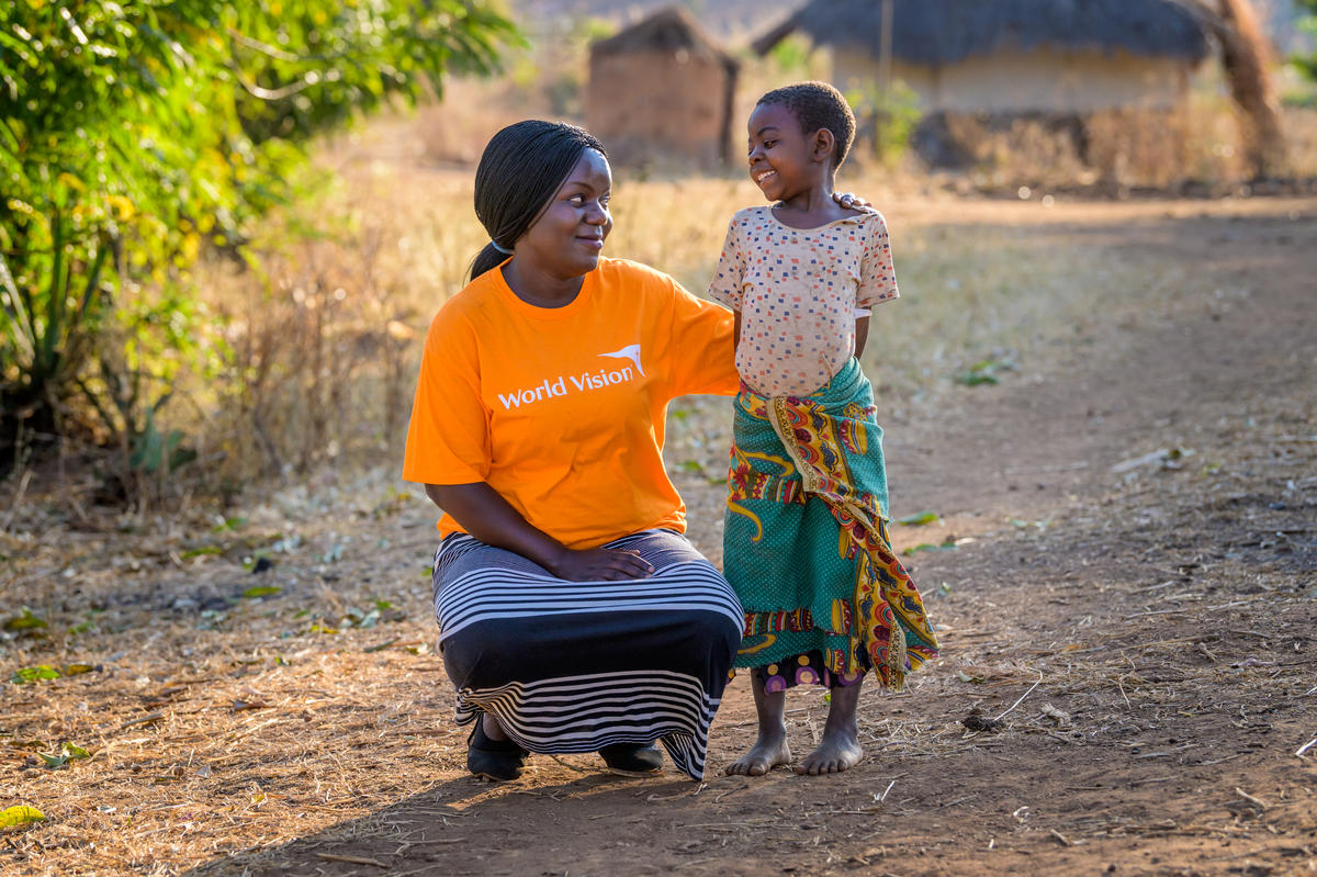 Trabajadora de desarrollo comunitario de World Vision con una camiseta naranja se agacha y sonríe y una niña que le devuelve la sonrisa en Malawi