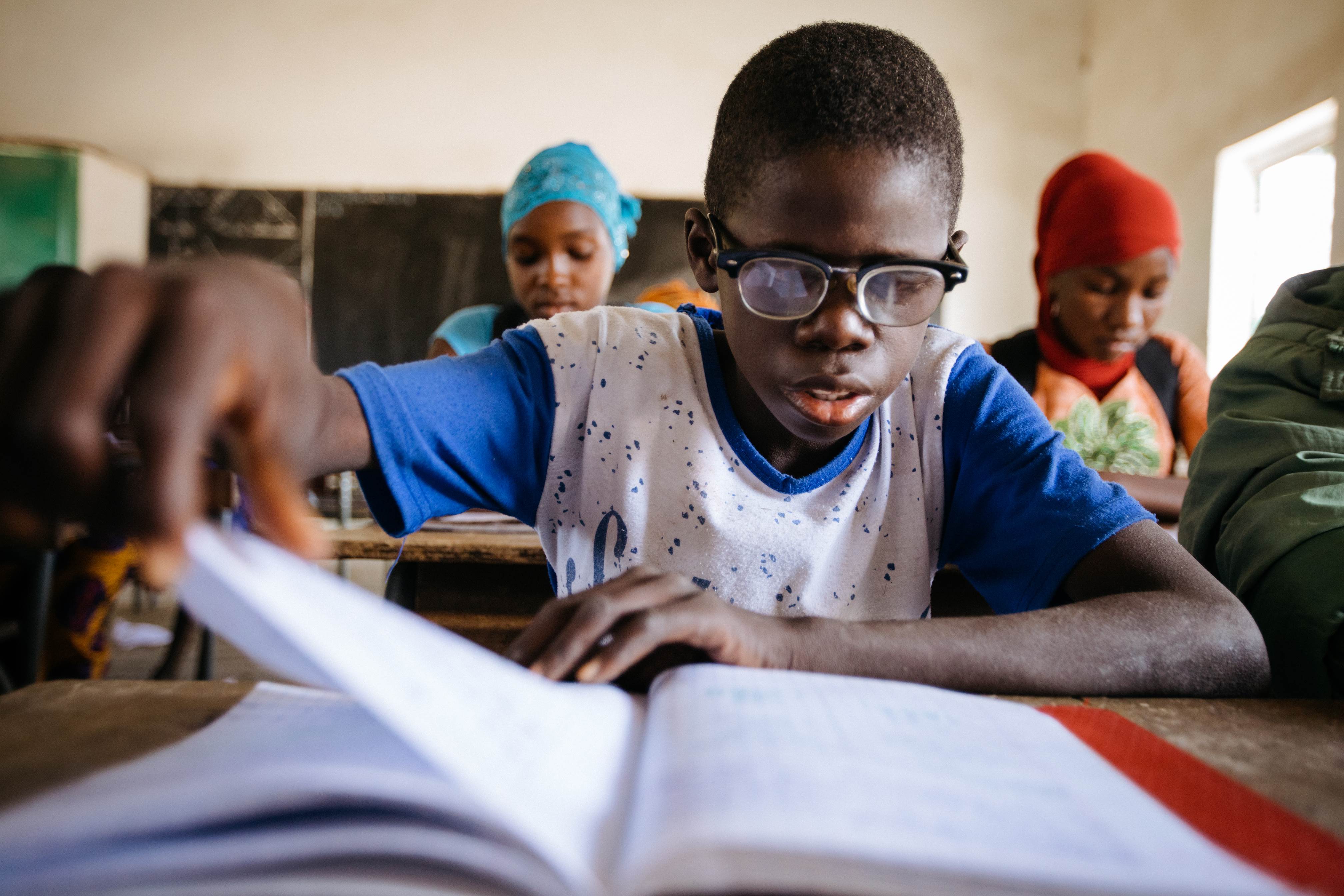 Un niño senegalés con gafas lee activamente un libro de texto en su pupitre de la escuela.