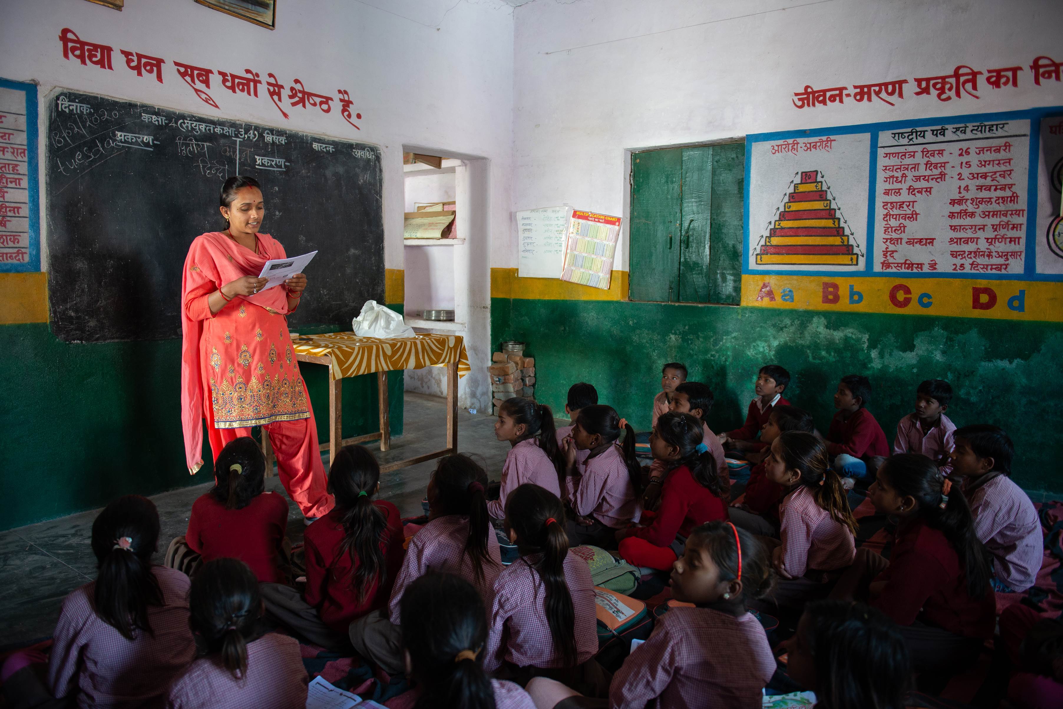 Una profesora vestida con un sari naranja brillante, dando clase a un grupo de niños en la India.