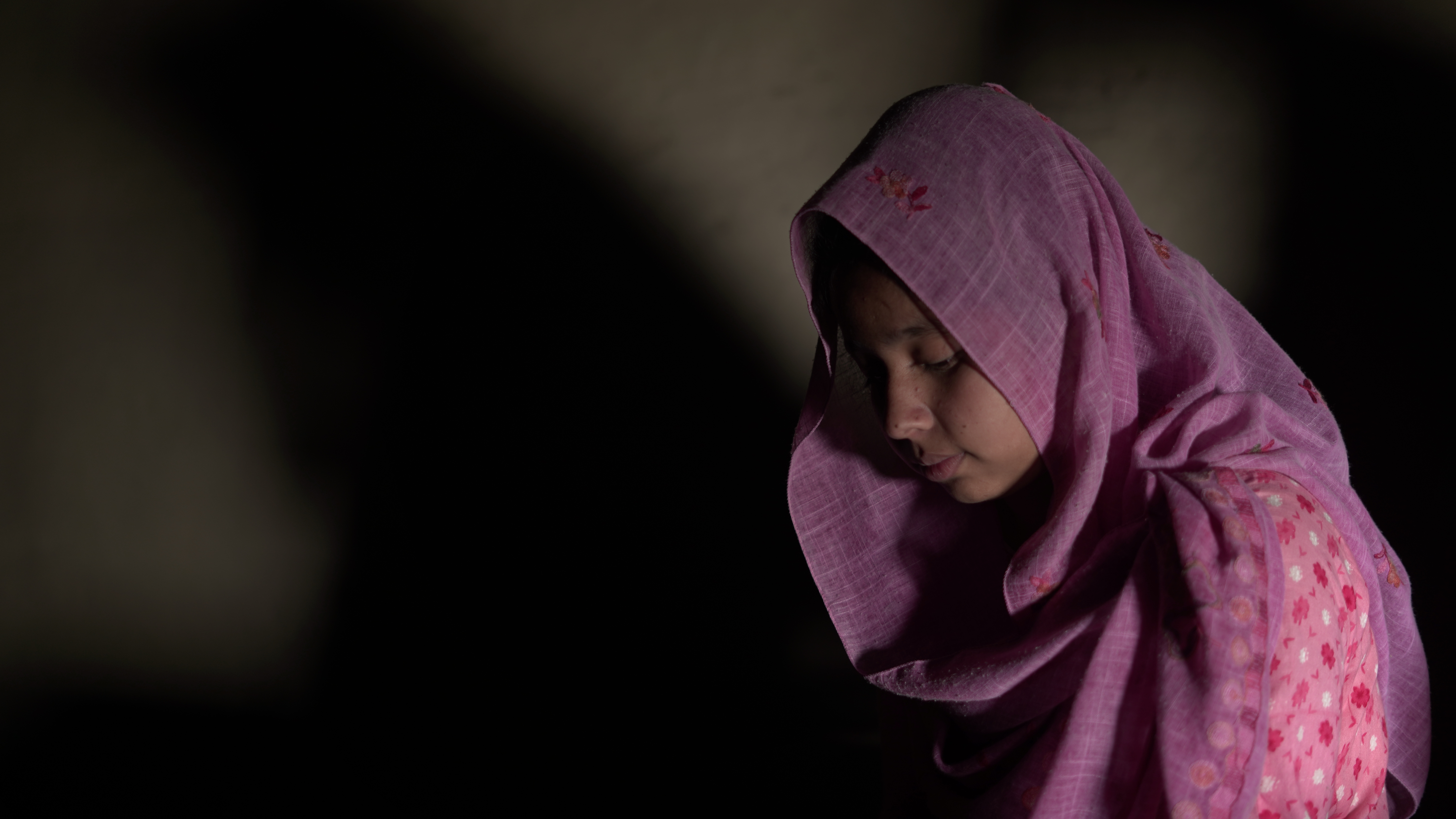 Niña bangladeshí con un pañuelo rosa brillante en la cabeza, de perfil, mirando hacia abajo en una habitación sucia.