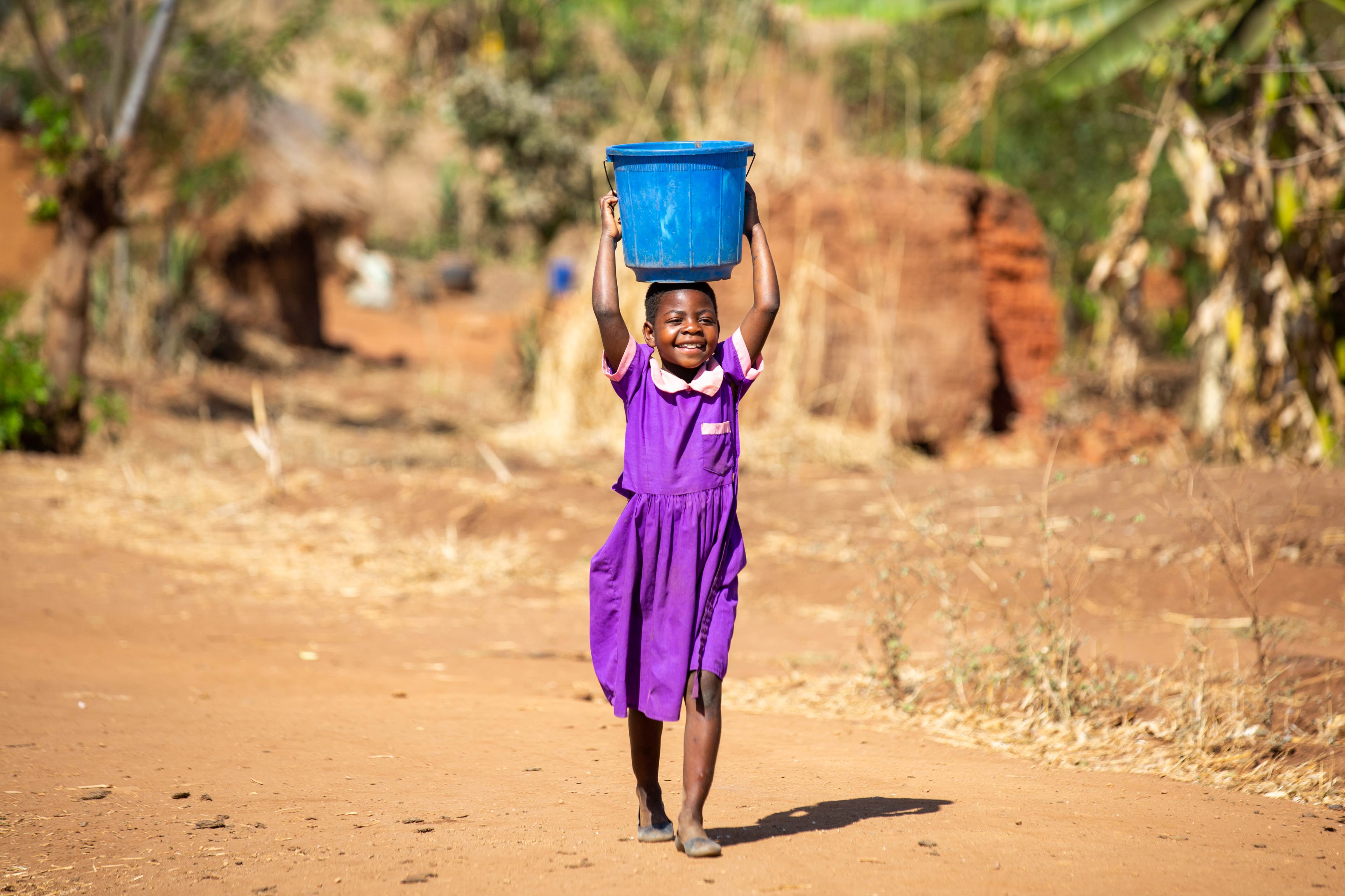Niña de 8 años de Malawi con un vestido púrpura brillante que lleva una gran cuba de agua potable en la cabeza.