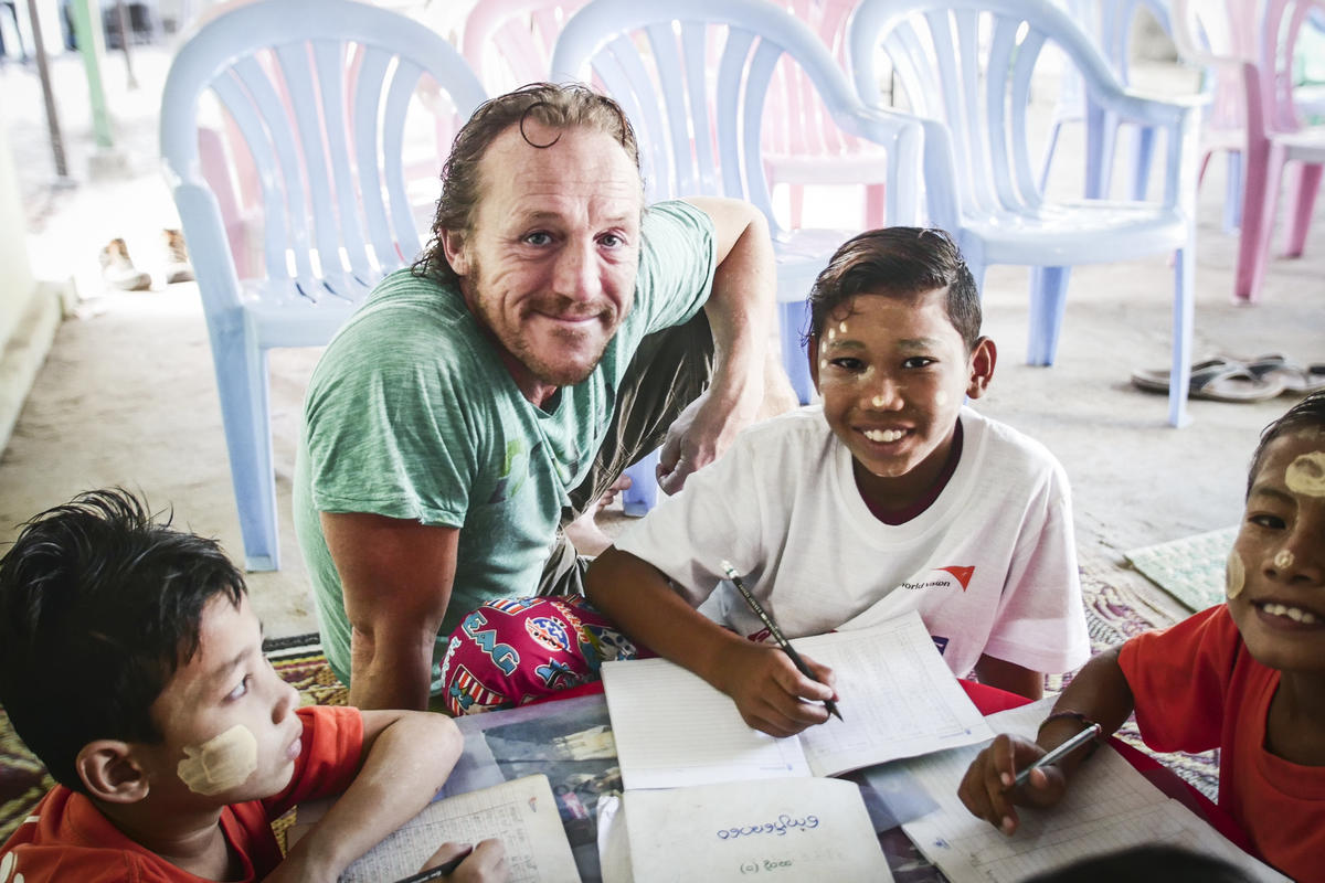 Jerome Flynn se sienta con un niño en Myanmar, el niño está trabajando en un cuaderno con un bolígrafo mientras Jerome sonríe